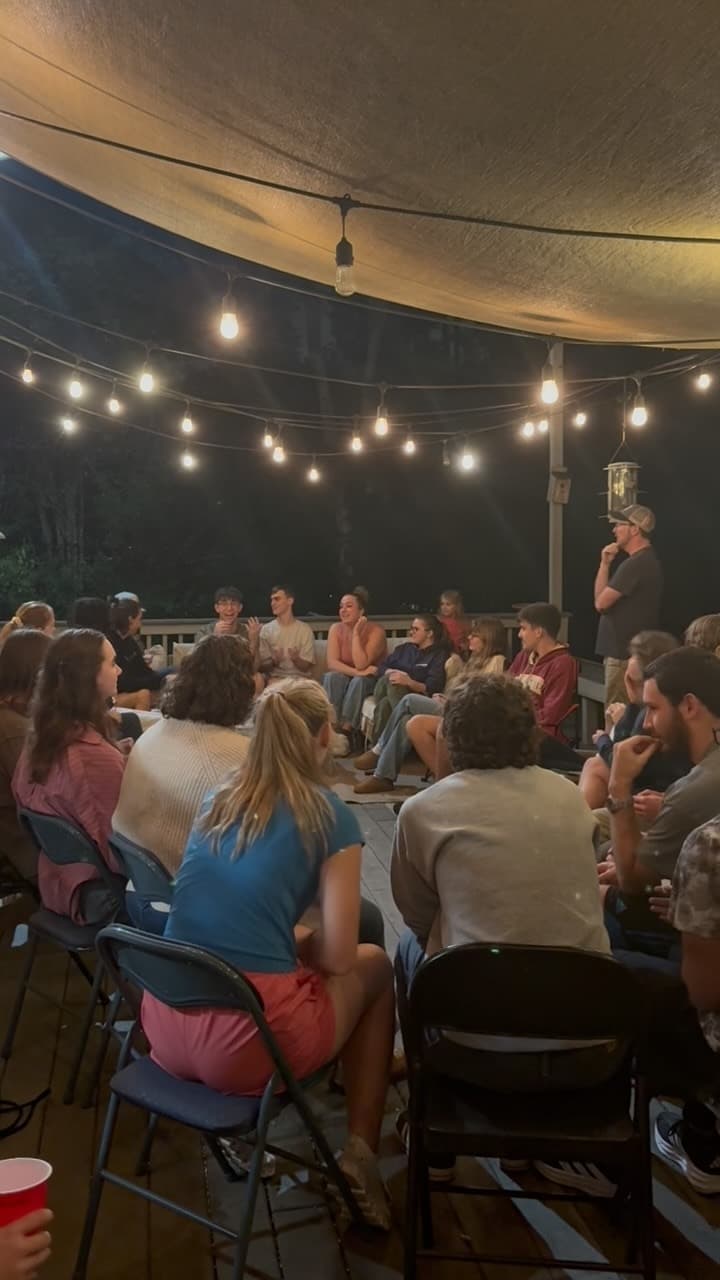 Students sitting together outdoors under string lights, listening to someone speak